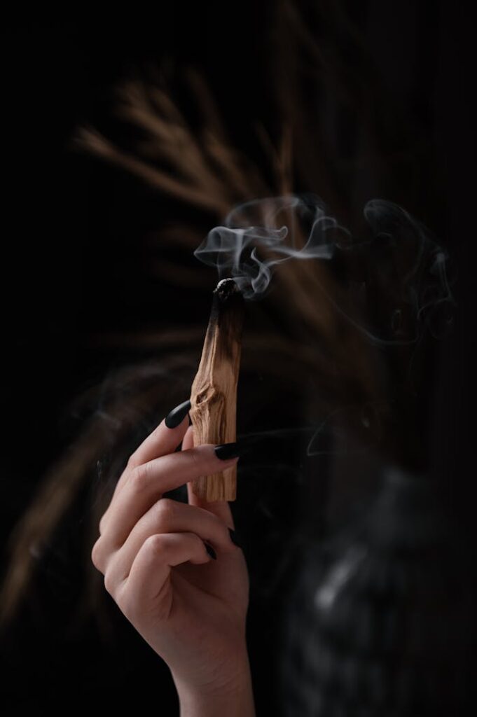 A hand holds burning palo santo stick, smoke rising in a dark, moody atmosphere.